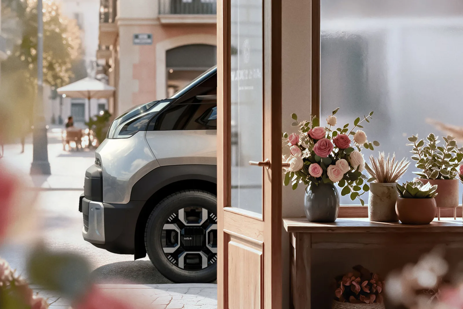 Kia PV7 i lys lak set forfra gennem en åben butiksdør på en solbeskinnet gade. Teaserbillede fra Kia.